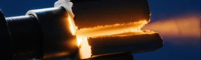 A metal test specimen clamped in a threaded grip, split by a jagged horizontal fracture that glows warm amber along the break, against a deep blue background.