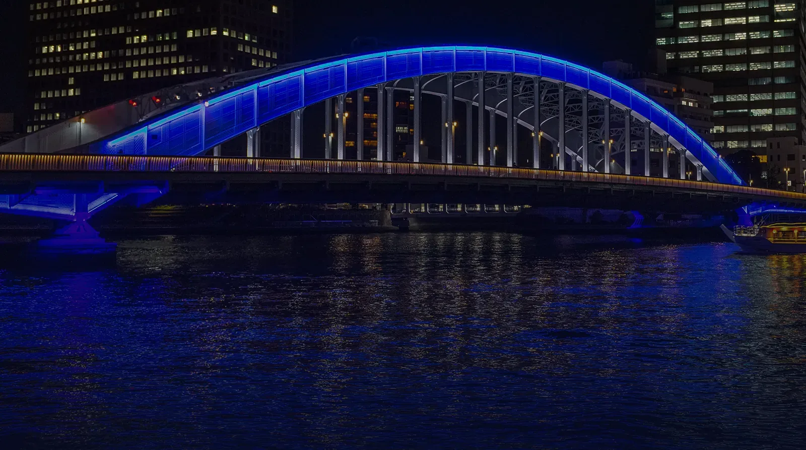 Illuminated steel arch bridge at night spanning a city river, reflected in the water