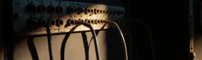 A worn switchboard panel of brass jack sockets, some with cables plugged in and others empty, lit by warm low light against a dark background.