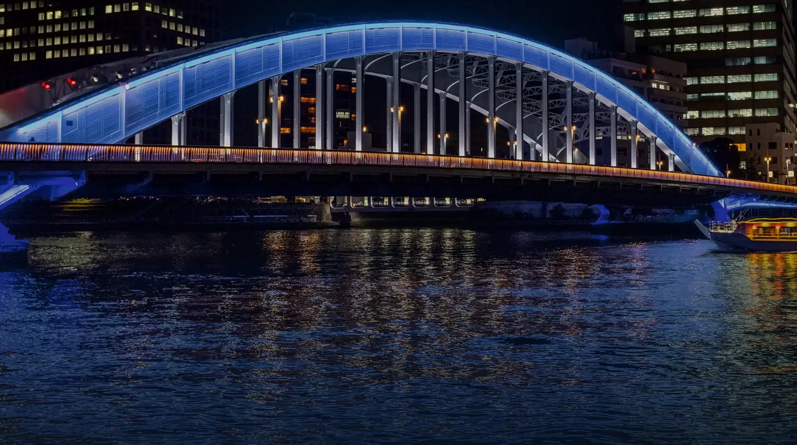 Illuminated steel arch bridge at night spanning a city river, reflected in the water